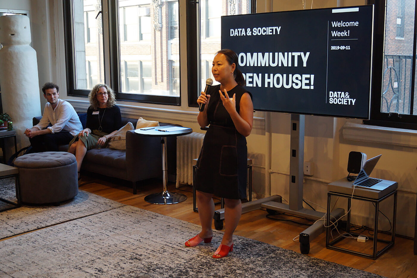 Woman with microphone standing in front of screen that reads Community Open House