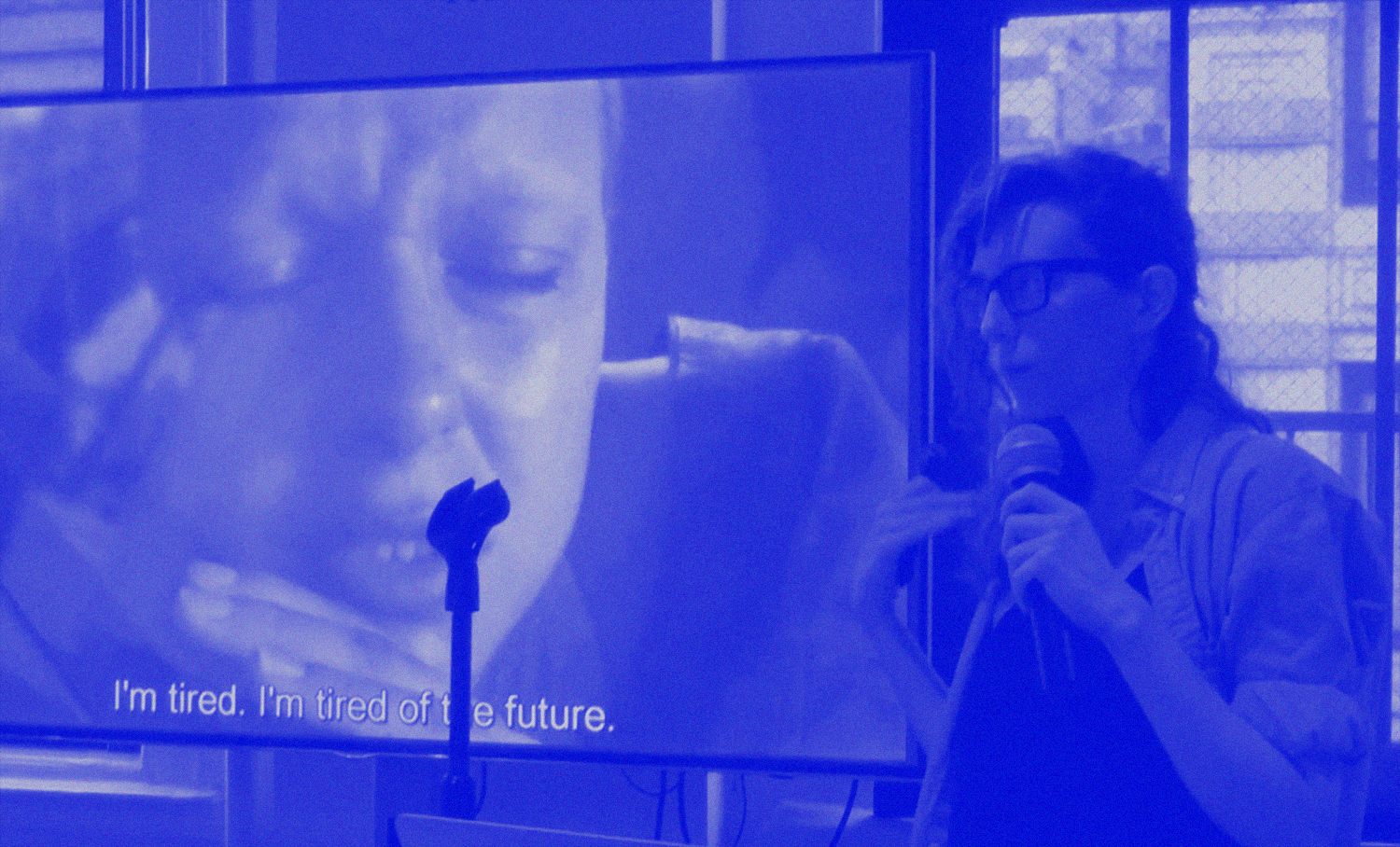photo of person speaking into microphone in front of movie screen-shot on monitor with a subtitle that reads, 