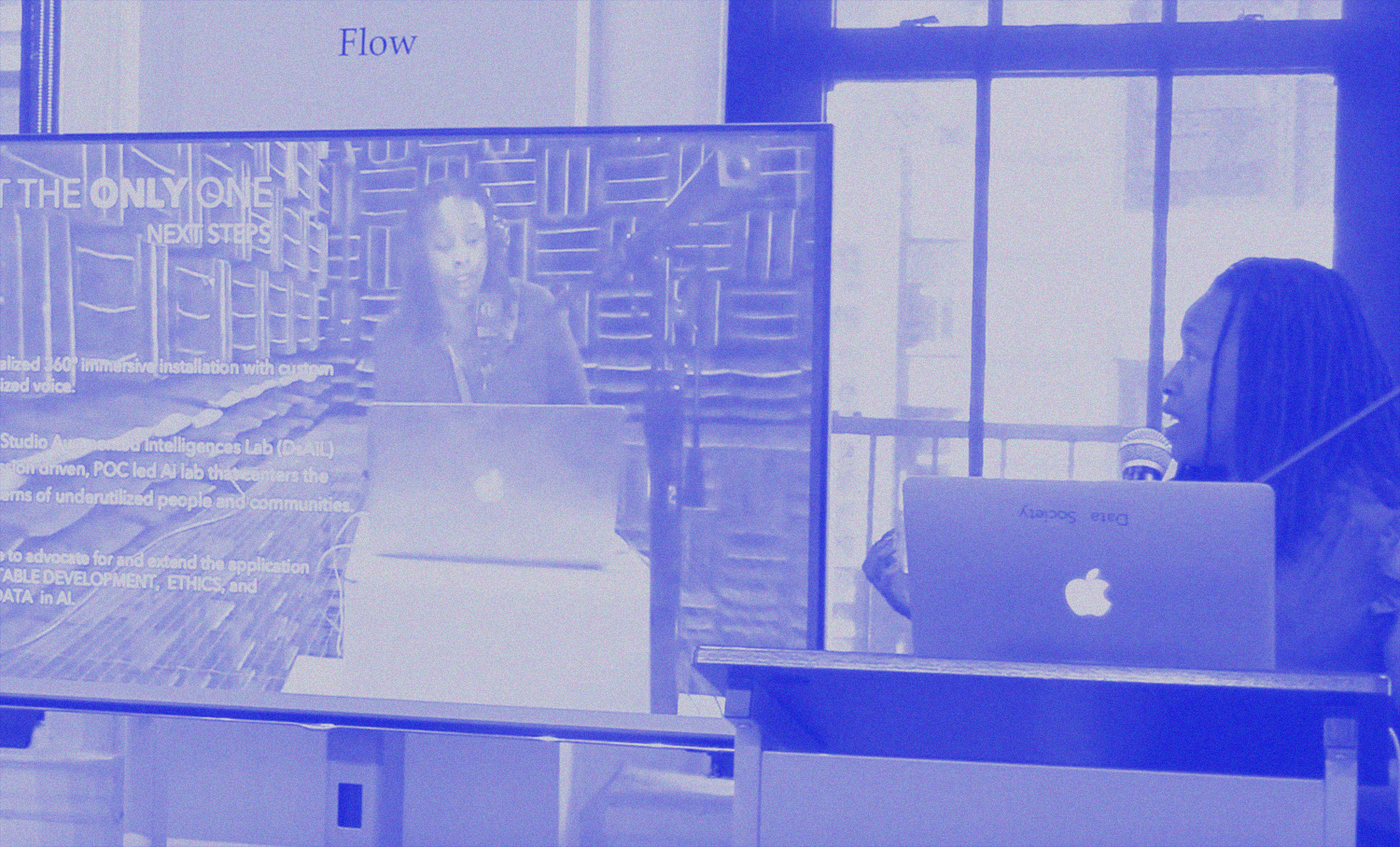 Photo of person speaking into microphone while looking at a screenshot of herself on the screen next to her.