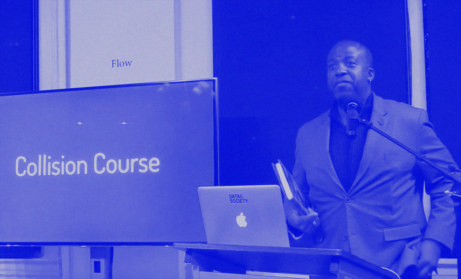 photo of man wearing a blazer, holding a book, and speaking next to a screen that says 